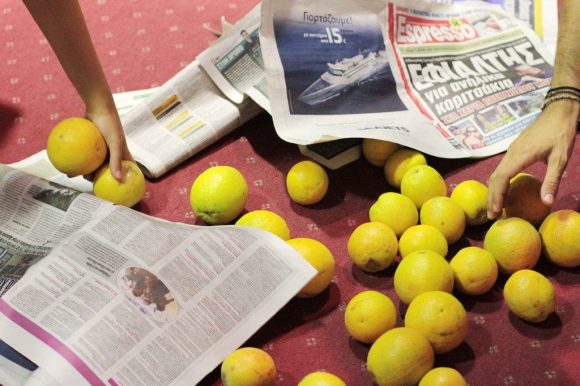 Πάνω σε μία κόκκινη μοκέτα είναι απλωμένα λεμόνια και φύλλα εφημερίδας. Δύο χέρια τείνουν να πάρουν μερικά από τα λεμόνια στο πάτωμα.